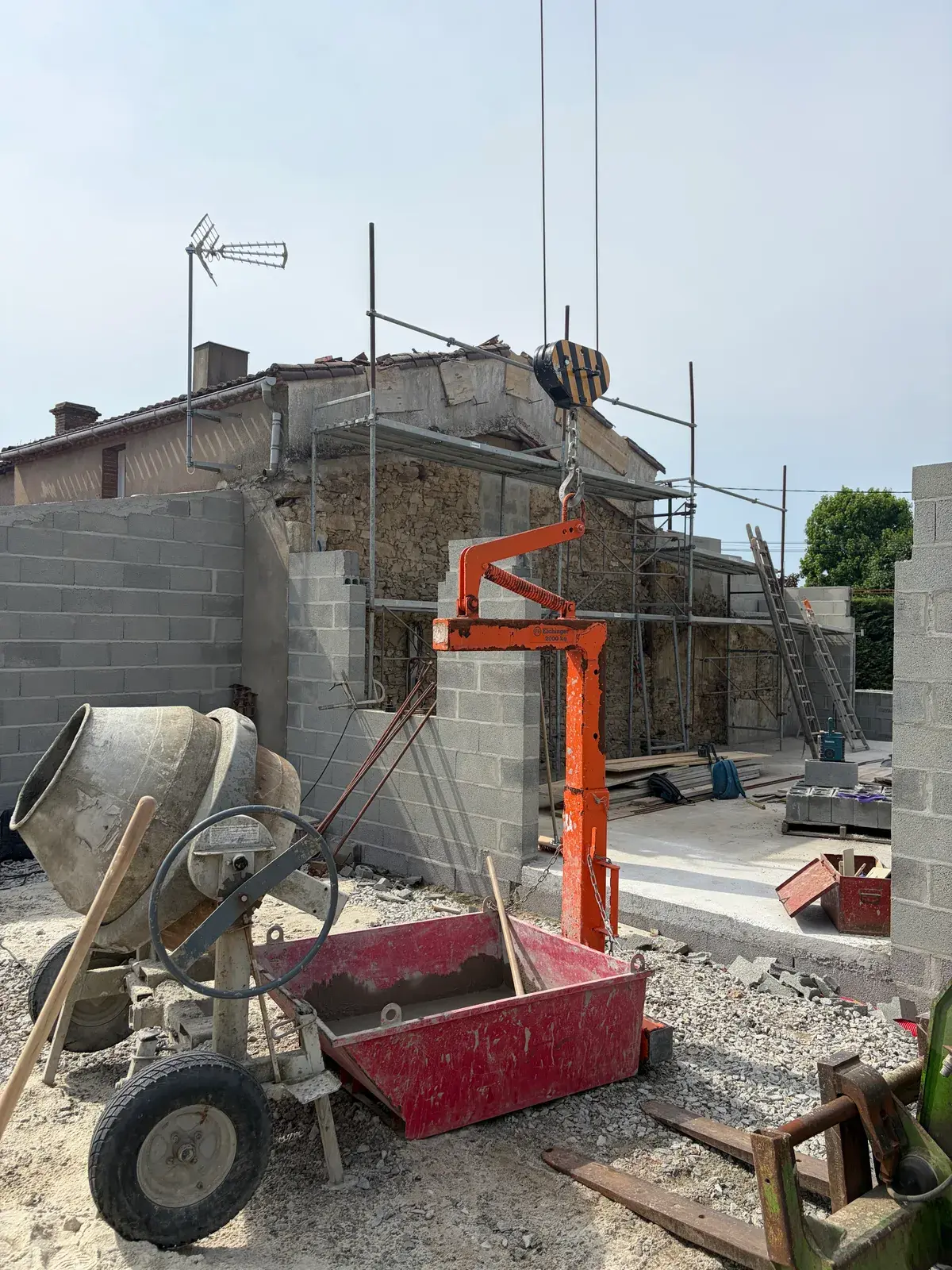 Ancien pignon en pierre en cours de déconstruction derrière les murs neufs en parpaings, benne à béton suspendue à la grue